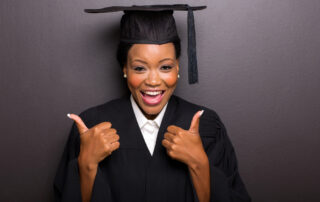 An adult college graduate in cap and gown smiles at the camera while giving a double thumbs-up.