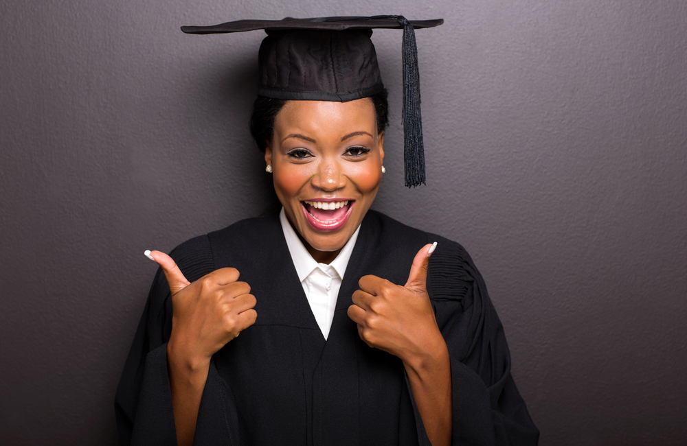 An adult college graduate in cap and gown smiles at the camera while giving a double thumbs-up.