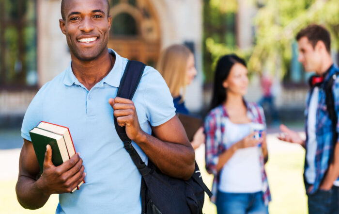 A smiling college student stands outside campus holding his books and a backpack.