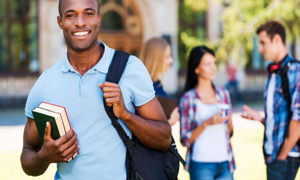 A smiling college student stands outside campus holding his books and a backpack.