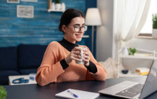 A smiling adult student enjoys a coffee while watching a lecture on her laptop.