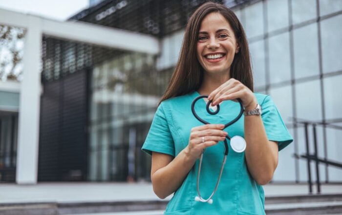 A smiling nurse forms a heart shape with her stethoscope.