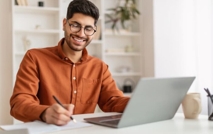 A smiling adult student taking notes while studying with his laptop